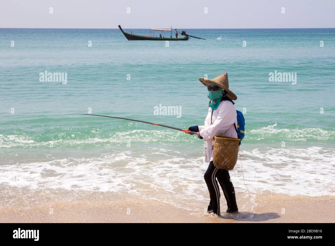 Phuket, Thailand-16. Oktober 2014: Eine Frau fischt vom Bang Tao Strand, wenn ein langes, verhülltes Boot vorbeiführt. Das Angeln ist ein Hauptstütze für einen Großteil des Phuket'schen Blocks Stockfoto