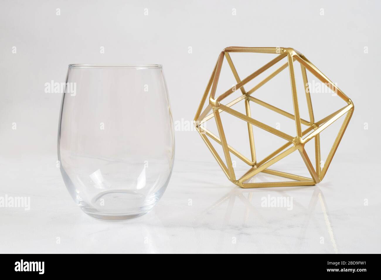 Ein luxuriöses, stielloses Weinglas schillt auf weißem Marmorhintergrund neben einer modernen Goldplastik. Viel Platz zum Kopieren, um Ihr eigenes Design hinzuzufügen Stockfoto