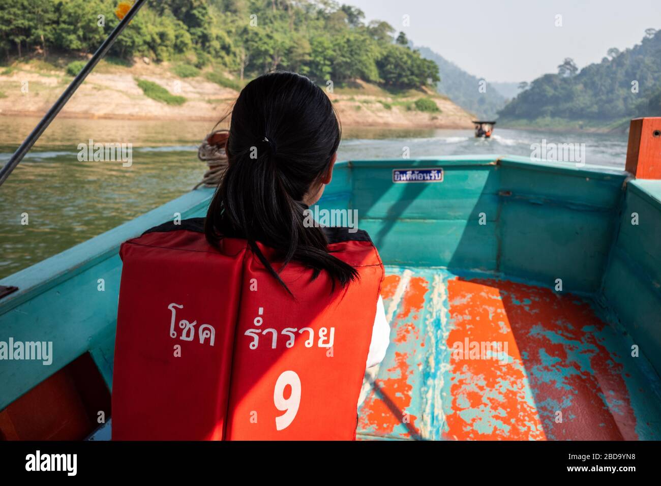 Nakhon Nayok, Thailand - 2. Februar 2020: Junges thailändisches Mädchen trägt orangefarbene Rettungsweste mit Namen des Bootes in thailändischer Sprache, während es Holzboot mit sp nimmt Stockfoto