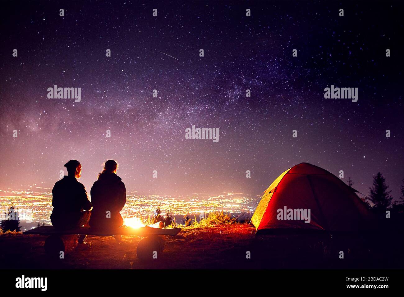 Glückliches Paar in Silhouette in der Nähe von Lagerfeuer und orange Zelt sitzen. Nachthimmel mit milchstrasse Sterne und die Lichter der Stadt im Hintergrund. Stockfoto