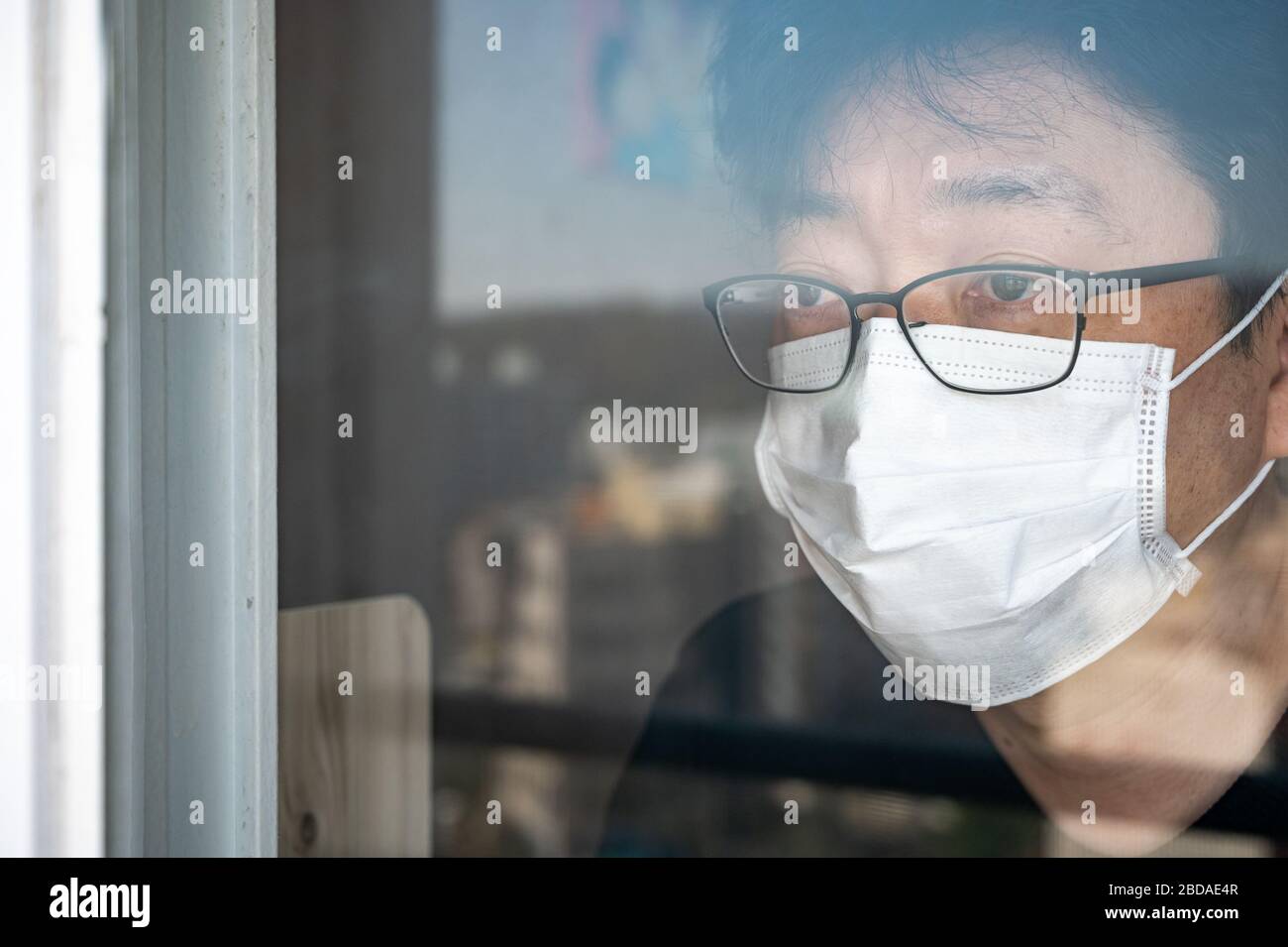 Selbstisolierungskonzept COVID-19 Coronavirus. Ein asiatischer Mann mit Gesichtsmaske und einem Fenster, das das Fenster während seiner Selbstisolation zu Hause auschaut. Stockfoto