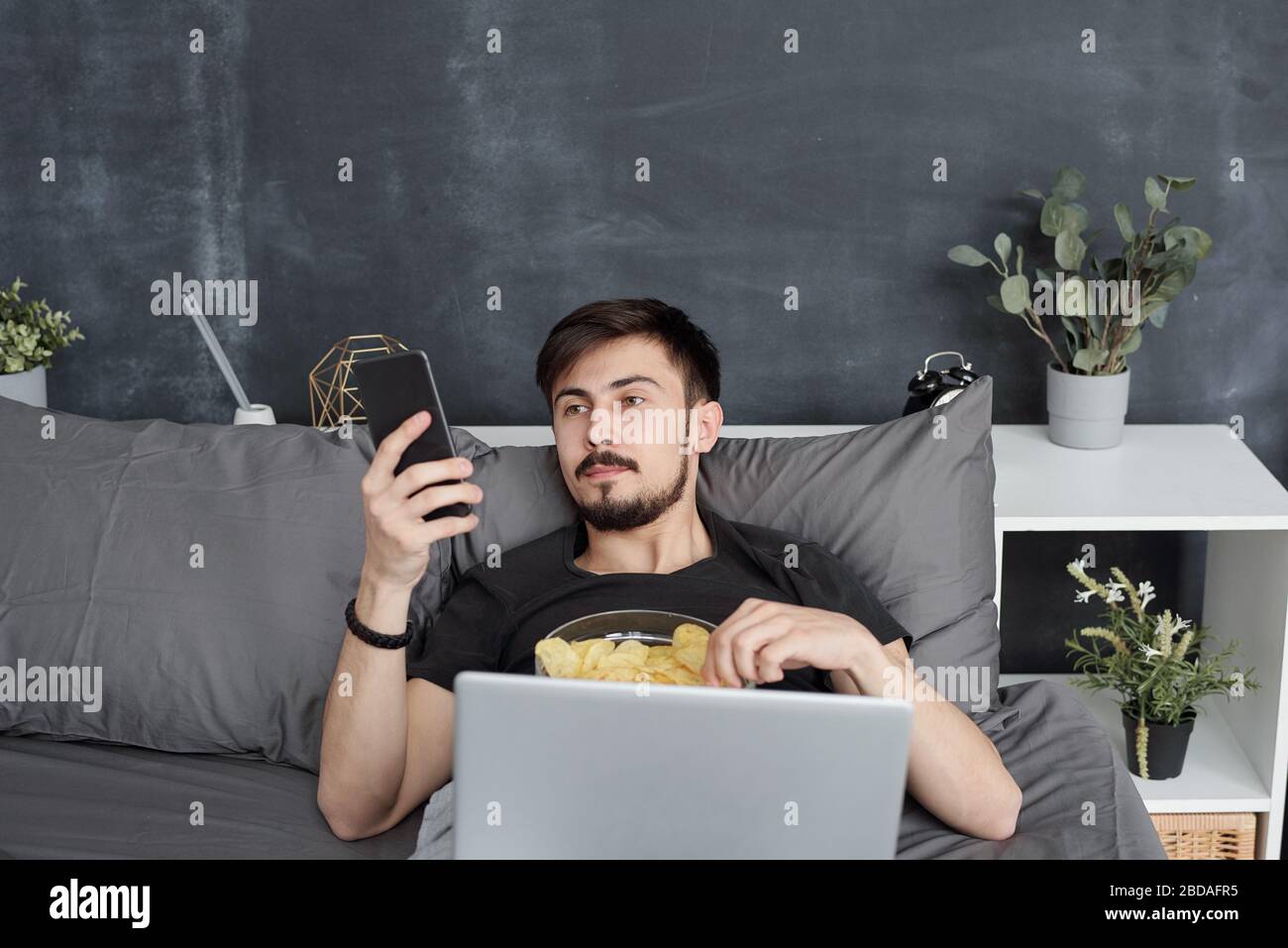 Langweiliger junger Student, der im Bett liegt und Chips isst, während er im Internet auf dem Smartphone surft, verschwendet er zu Hause Zeit während der Quarantäne Stockfoto