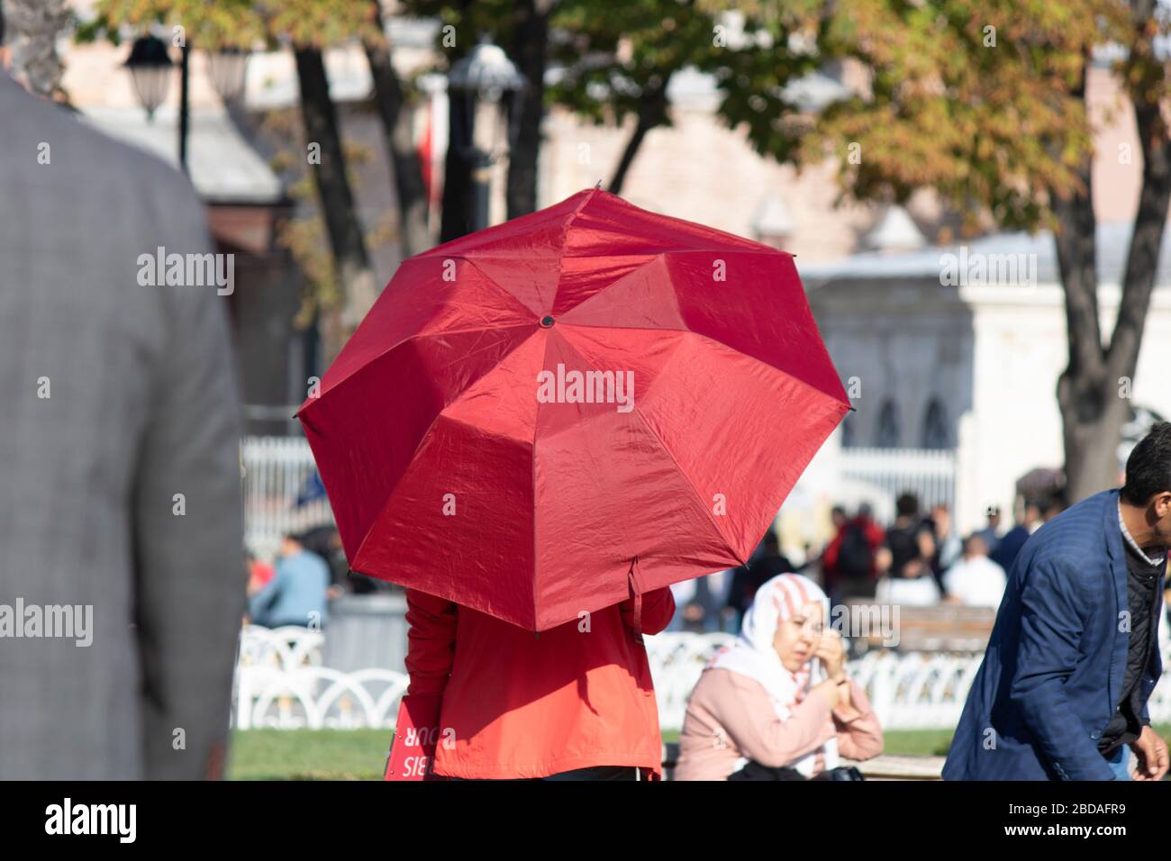 Dame, die mit rotem Regenschirm auf dem Platz spaziert. Er ist vor der Sonne geschützt. Touristen gehen um. Ein Foto wurde auf dem Sultanahmet-Platz gemacht. Stockfoto