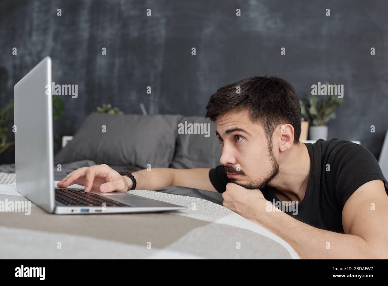 Langweiliger junger bärtiger Mann, der auf dem Laptop-Bildschirm schaut, während er auf Websites in Quarantäne surft Stockfoto