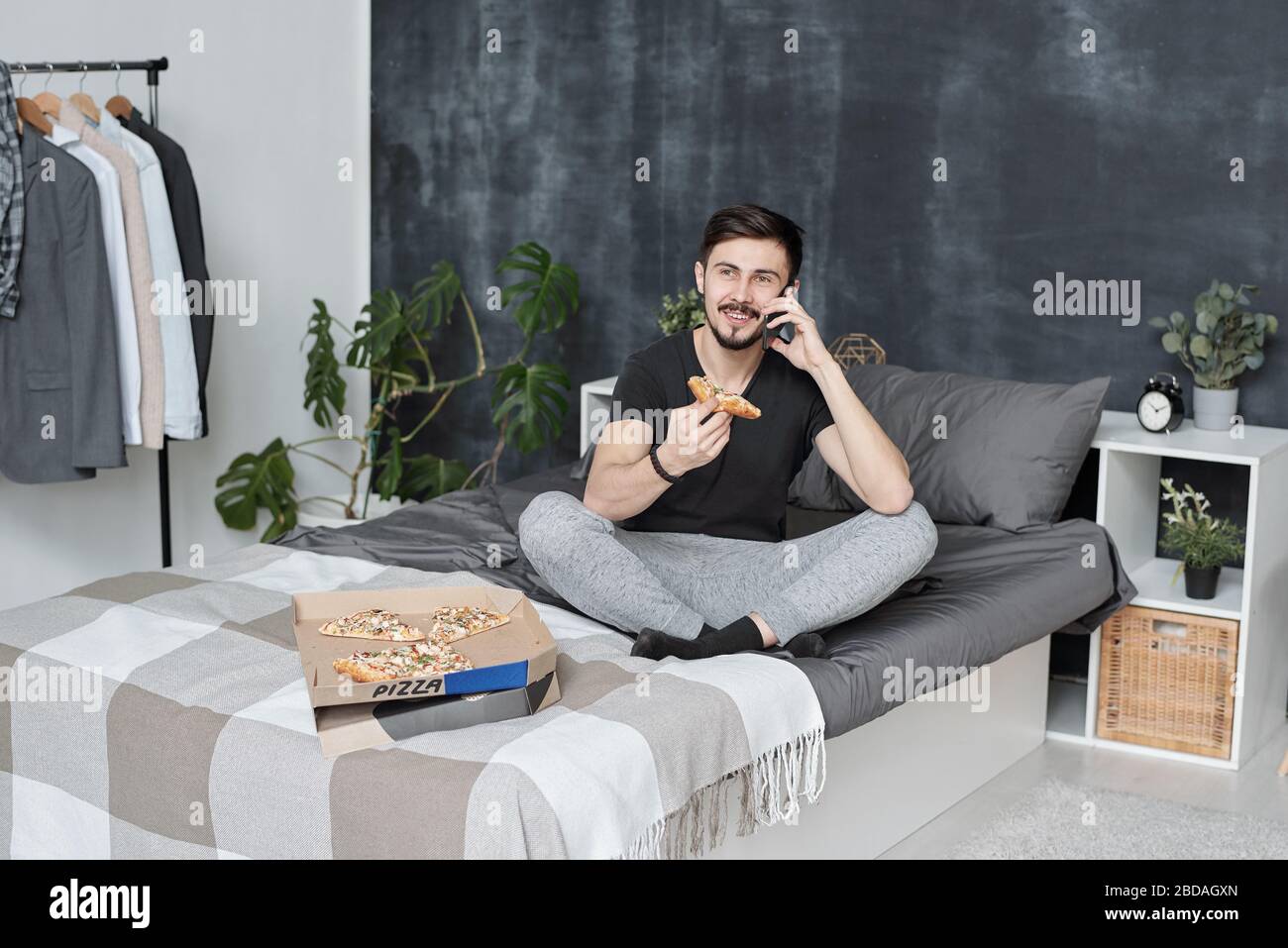Fröhlicher, gutaussehender junger bärtiger Mann, der auf dem Bett sitzt und telefoniert, während er zu Hause Pizza isst Stockfoto