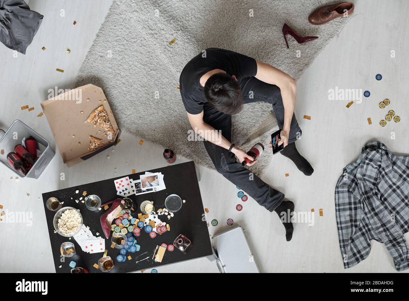 Über dem Blick auf den brünettigen Mann, der auf dem Boden sitzt und unordentliche Casino-Chips und Snacks hat und Partyfotos auf dem Smartphone sieht, während er morgens Bier trinkt Stockfoto