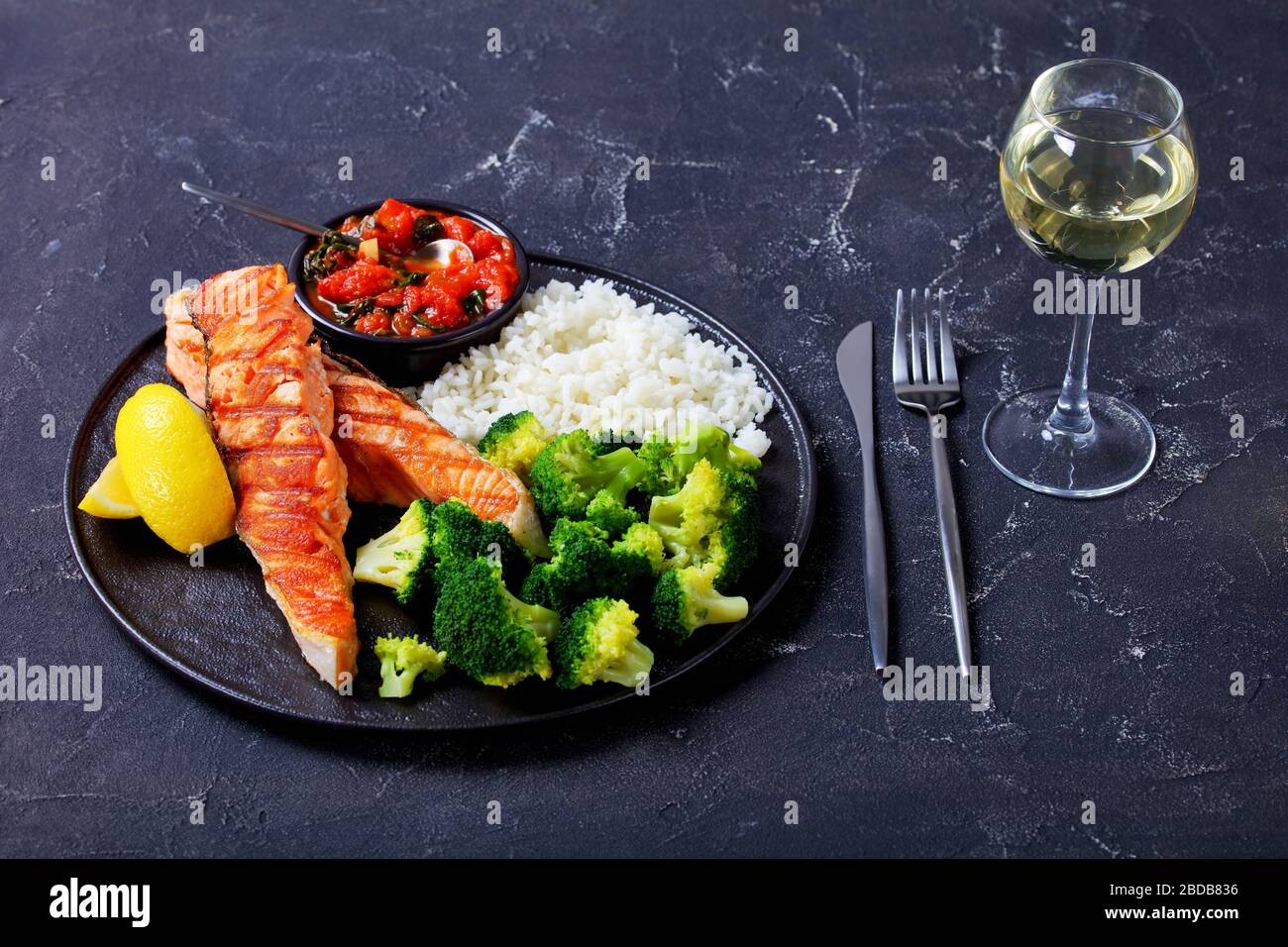 Platte aus gegrilltem Lachssteak mit gedämpftem Brokkoli, Langkornreis, Tomaten- und Spinatsoße, Zitronenkeile auf einem dunklen Betontisch mit einem Glas Stockfoto