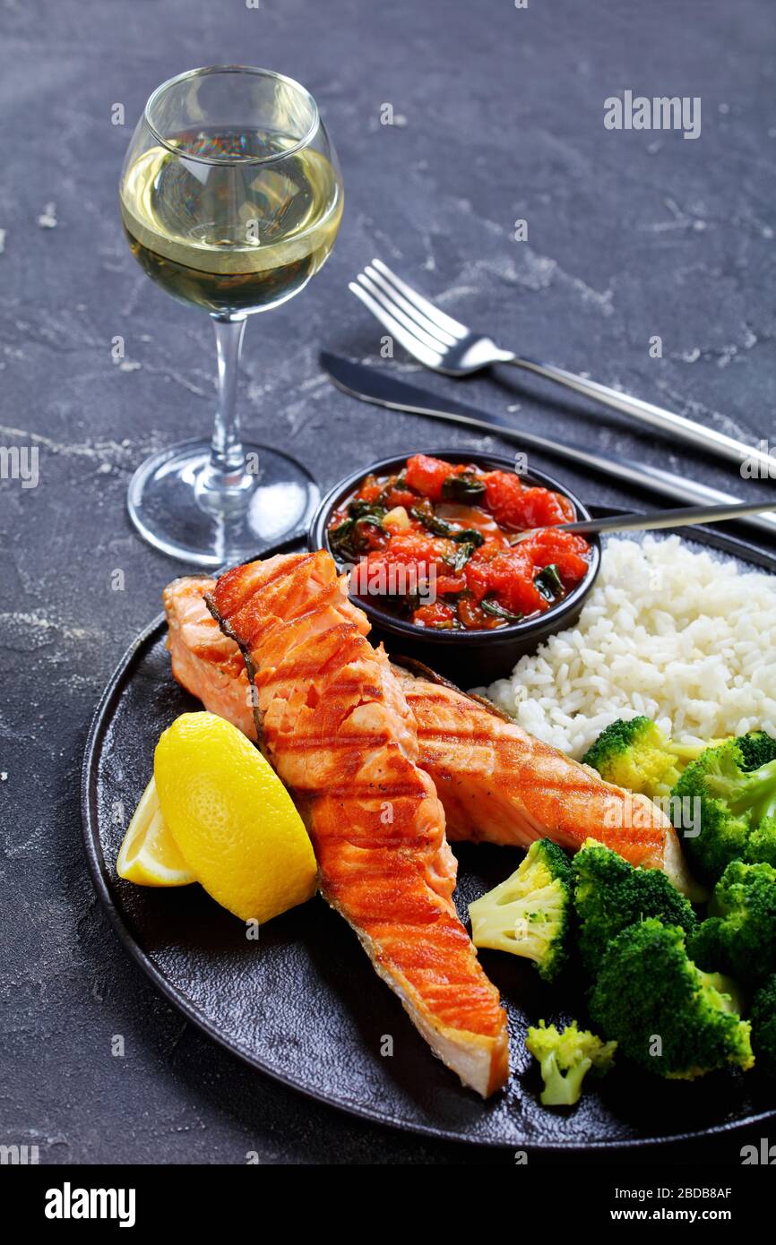 Restaurant serviert: Lachssteak mit Gemüse: Brokkoli, Tomate und Spinatsoße und Langkornreis, Zitronenkeile auf einer schwarzen Platte mit Weiß Stockfoto