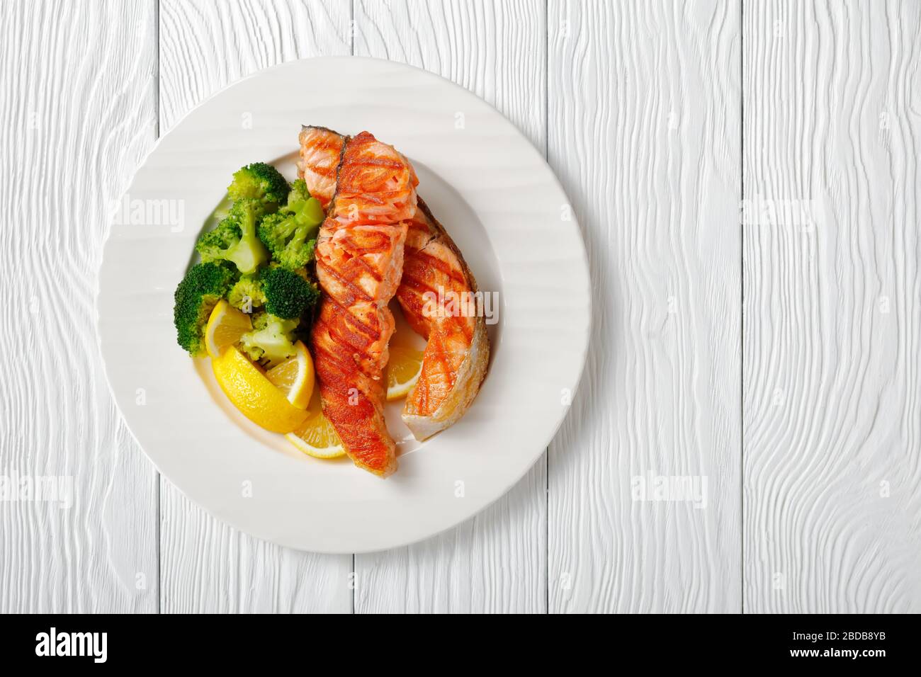 Gesundes Abendessen mit gegrilltem Lachs mit gekochten Brokkoli, Zitronenkeilen auf einem weißen Teller mit goldenem Besteck auf weißem Holzhintergrund, flacher Lay Stockfoto