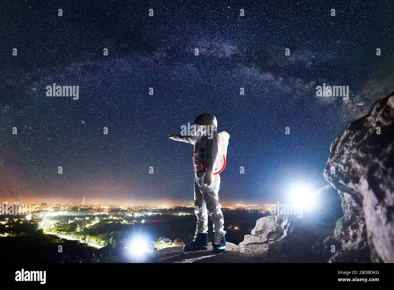 Männlicher Spaceman, der mit dem Finger auf die Stadt in der Nacht zeigt, während er auf felsigen Bergen unter dem fantastischen Nachthimmel mit Sternen und Milchstraße steht. Konzept der Kosmonauten und Raumfahrt. Stockfoto