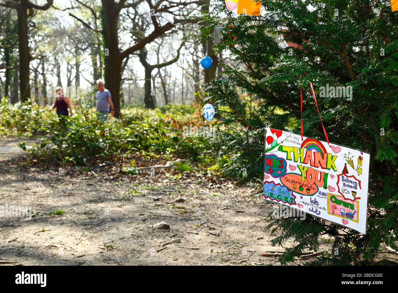 April 2020, Southborough Common, Kent, Großbritannien: Botschaften der Kinder, die NHS-Mitarbeitern und wichtigen Arbeitern danken, die an einem Baum hängen, der während der von der Regierung verhängten Quarantäne/Sperrung für Southborough Common gestellt wurde, um die Ausbreitung des Coronavirus zu verringern. Kinder im ganzen Land haben Botschaften und Zeichnungen von Regenbögen in Fenster und anderswo gesetzt, um Hoffnung zu verbreiten und die Menschen zu ermutigen, während der Pandemie fröhlich zu bleiben. Ein älteres Paar ist im Hintergrund zu sehen. Stockfoto