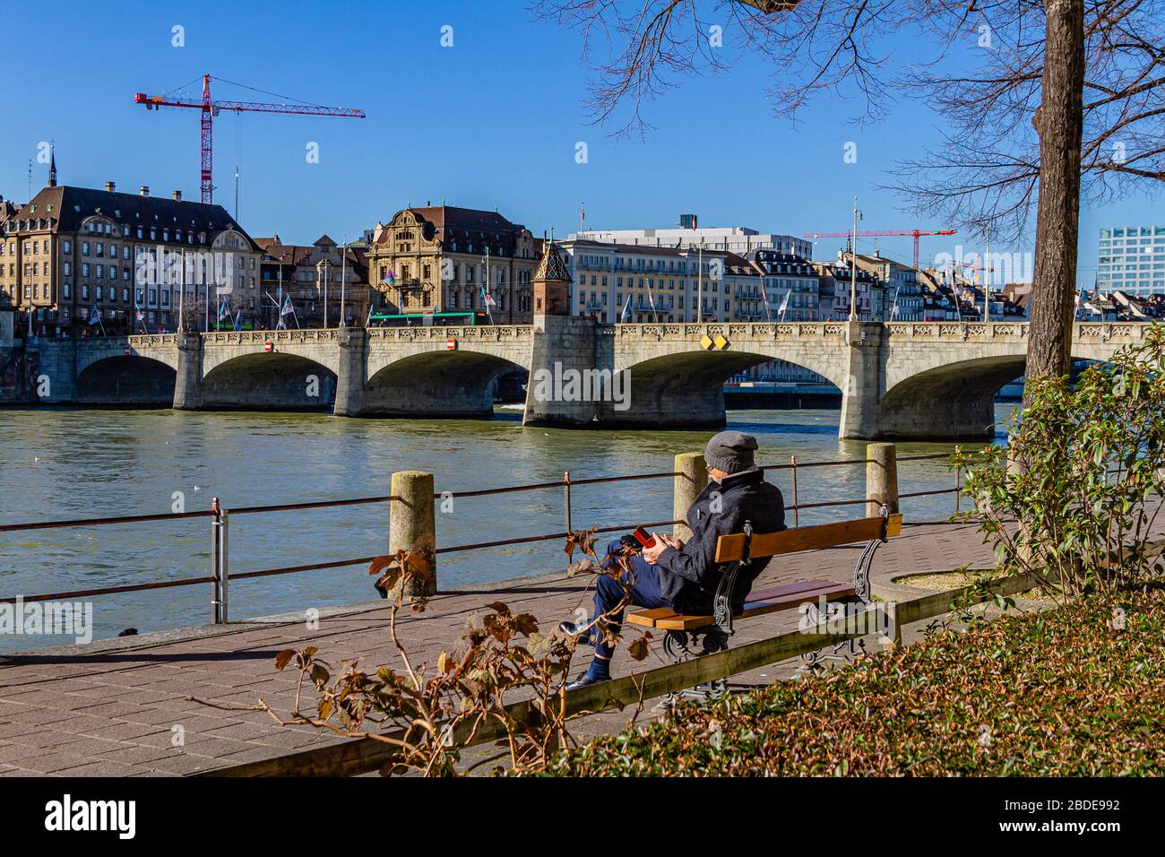 Eine Person, die auf einer Bank neben dem Rhein und der historischen Mittelbrücke in der Stadt Basel, Schweiz sitzt. Februar 2020. Stockfoto
