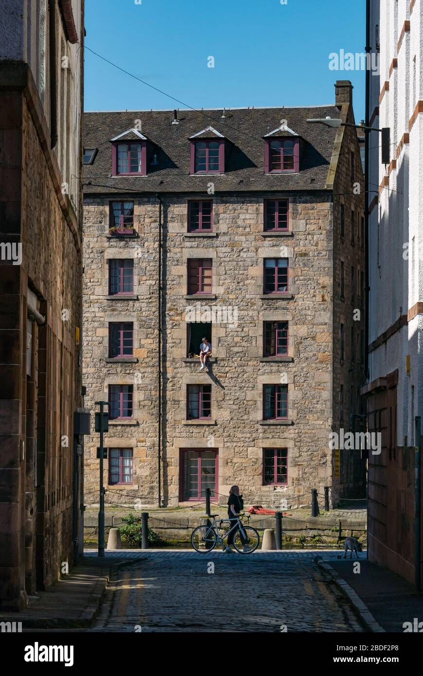 Leith, Edinburgh, Schottland, Großbritannien. April 2020. Covid-19 Lockdown: An einem der wärmsten Sonnentage in diesem Jahr mit Menschen in Lockdown. Ein Mann und sein Hund sitzen in einem offenen Fenster im 3. Stock des Cooperage-Lagers, das in Wohnungen am Ufer entlang des Leith-Wassers umgewandelt wurde, prekär Stockfoto