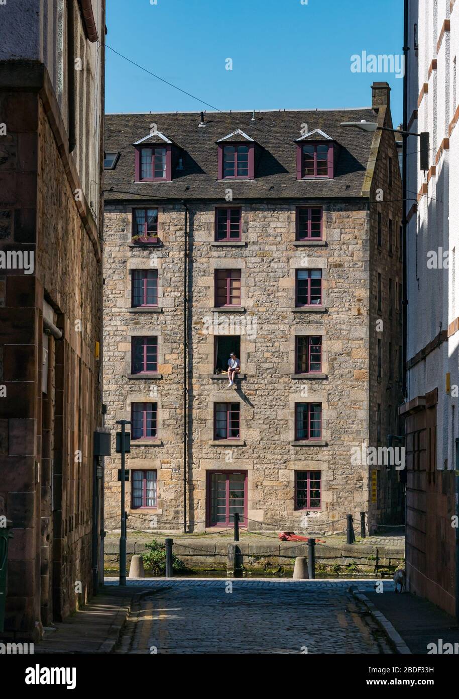 Leith, Edinburgh, Schottland, Großbritannien. April 2020. Covid-19 Lockdown: An einem der wärmsten Sonnentage in diesem Jahr mit Menschen in Lockdown. Ein Mann und sein Hund sitzen in einem offenen Fenster im 3. Stock des Cooperage-Lagers, das in Wohnungen am Ufer entlang des Leith-Wassers umgewandelt wurde, prekär Stockfoto