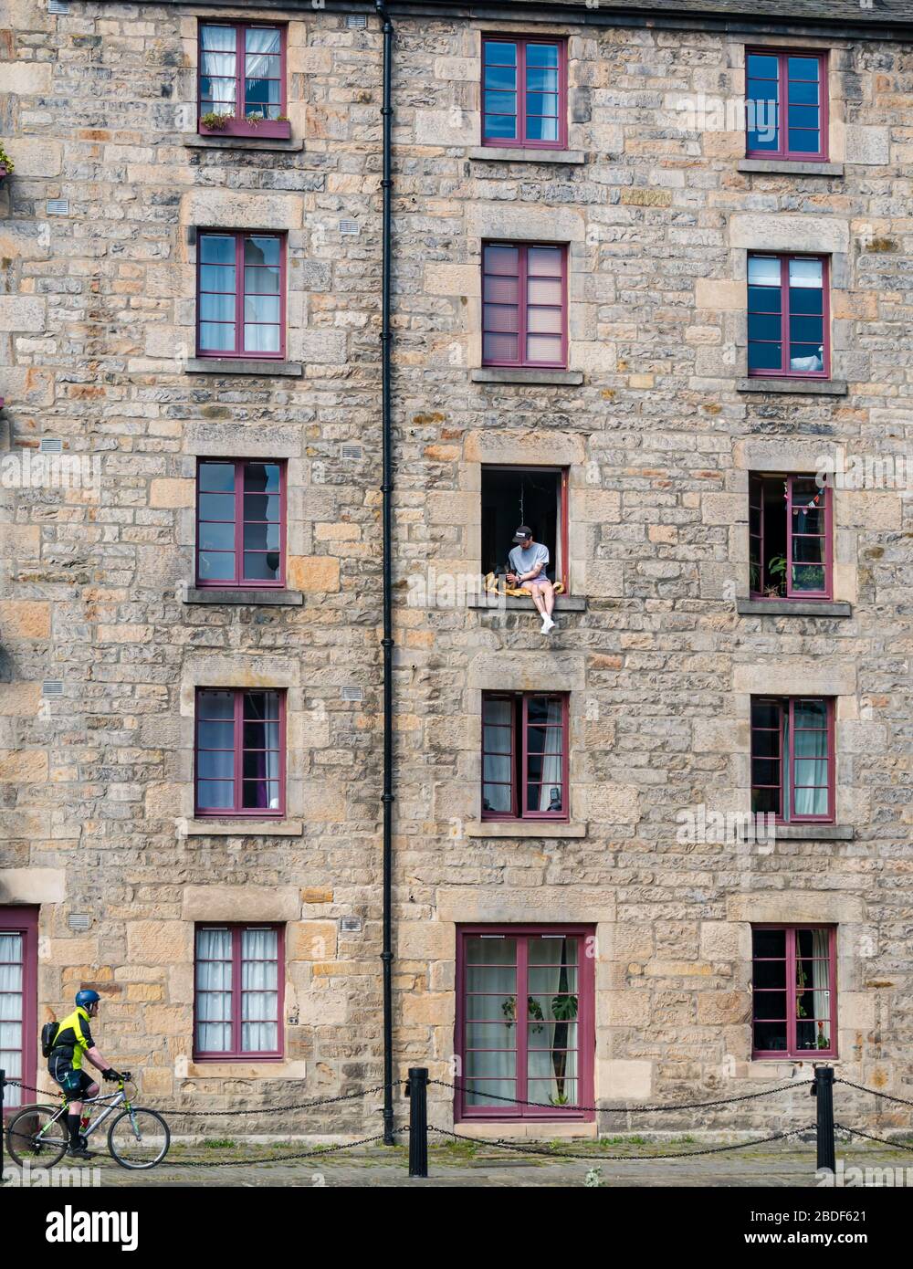 Leith, Edinburgh, Schottland, Großbritannien. April 2020. Covid-19 Lockdown: An einem der wärmsten Sonnentage in diesem Jahr mit Menschen in Lockdown. Ein Mann und sein Hund sitzen in einem offenen Fenster im 3. Stock des Cooperage-Lagers, das in Wohnungen am Ufer entlang des Leith-Wassers umgewandelt wurde, prekär Stockfoto