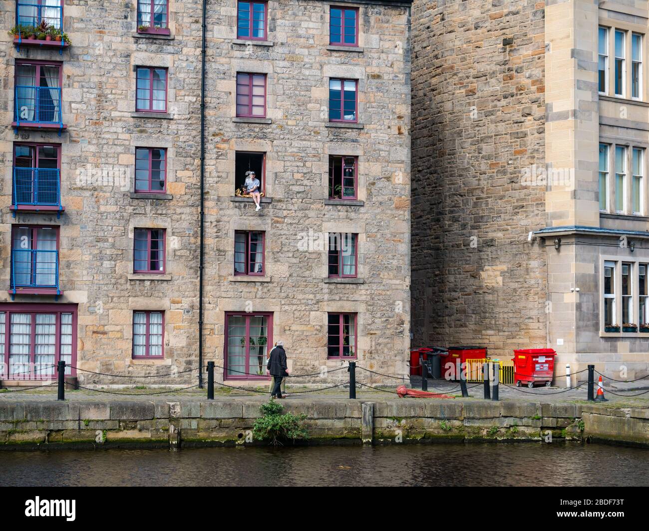 Leith, Edinburgh, Schottland, Großbritannien. April 2020. Covid-19 Lockdown: An einem der wärmsten Sonnentage in diesem Jahr mit Menschen in Lockdown. Ein Mann und sein Hund sitzen in einem offenen Fenster im 3. Stock des Cooperage-Lagers, das in Wohnungen am Ufer entlang des Leith-Wassers umgewandelt wurde, prekär Stockfoto