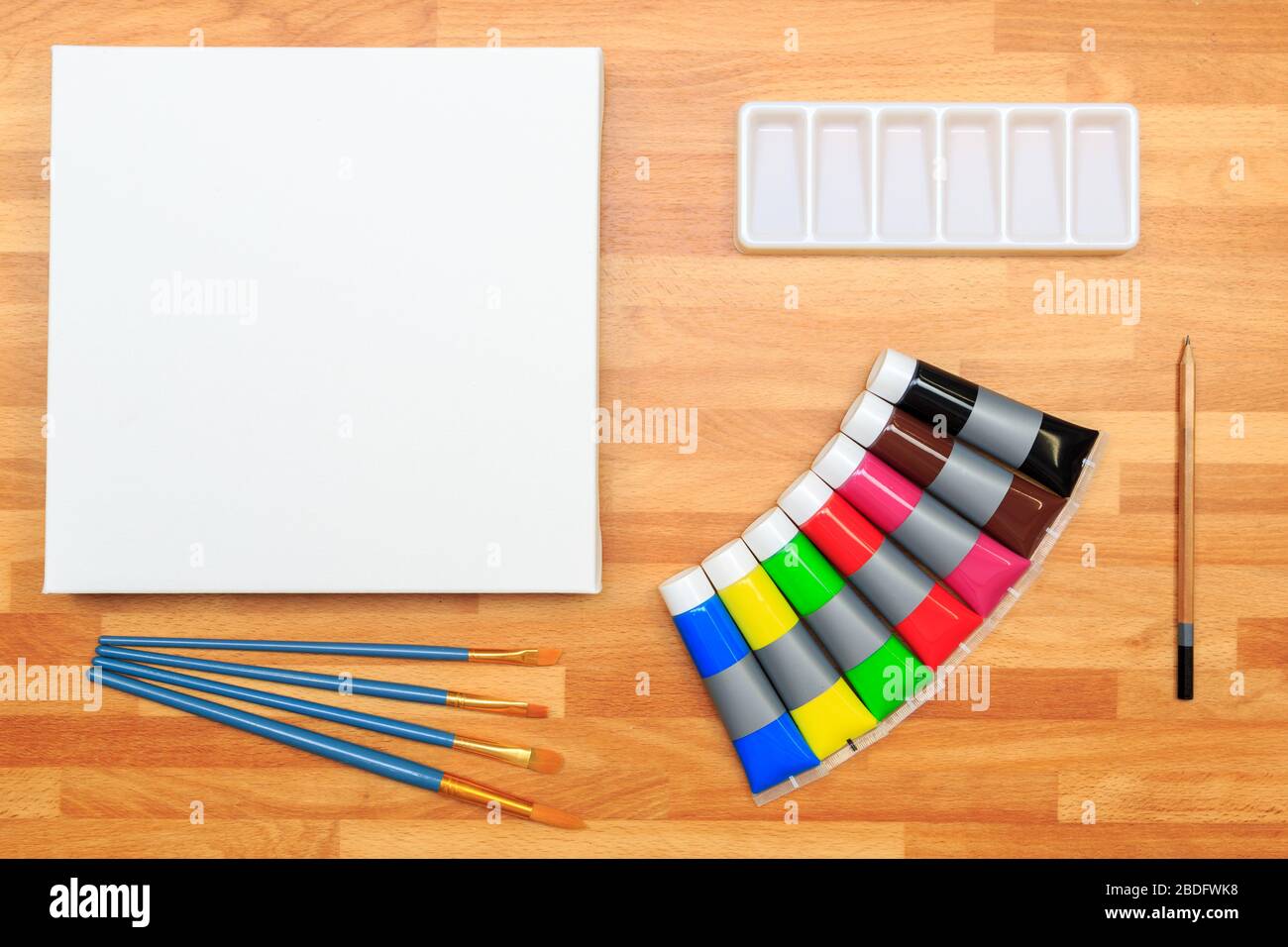 Blick auf Farbenröhrchen, Farbpalette, Bürsten, Bleistift und weiße Leinwand auf einem Holzhintergrund Stockfoto