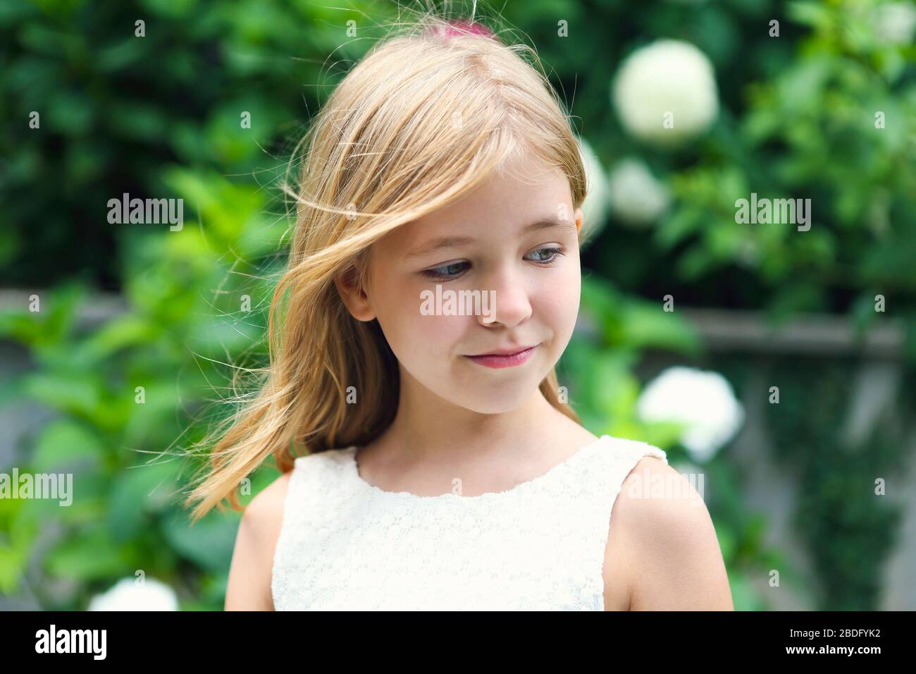 Fröhliches kleines Mädchen mit Ferkelschwanz in weißem Kleid lächelnd und mit Blick auf den grünen Garten am Sommerabend Stockfoto