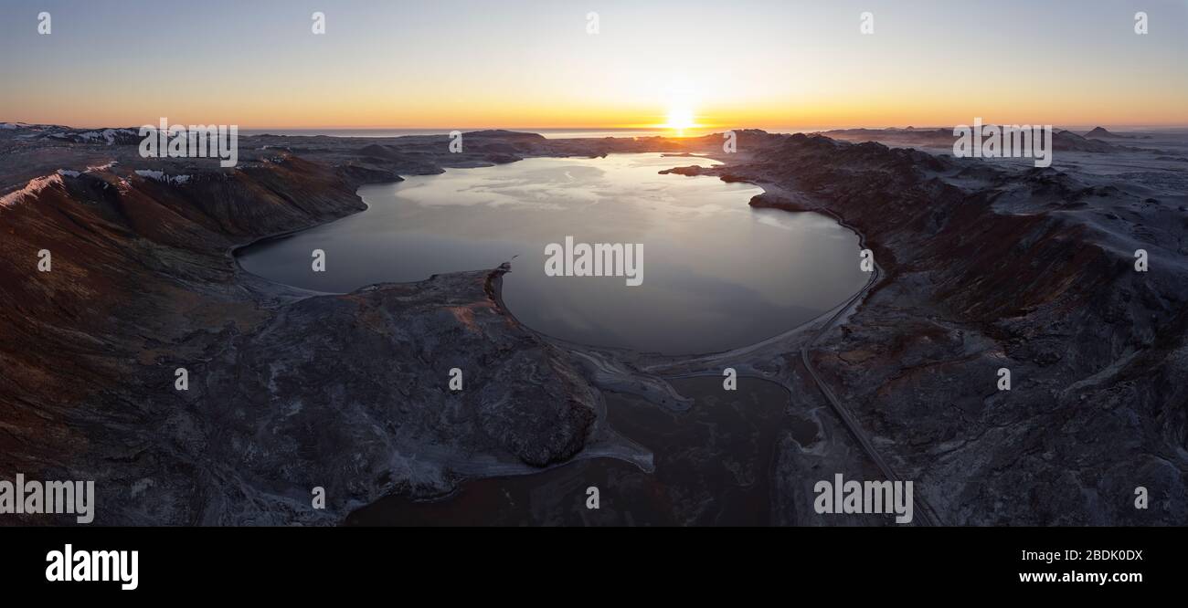 Spektakuläre nordische Panoramalandschaft mit ruhigem See, umgeben von unwegsamem, felsigem Gelände gegen klaren Himmel mit Sonne über Horizont in Island Stockfoto