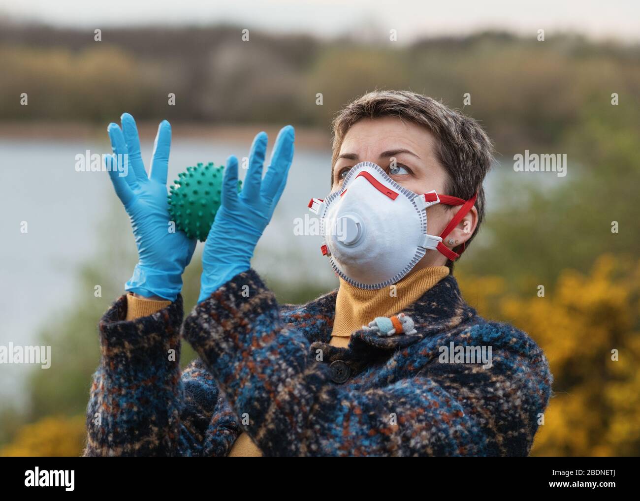 Weicher Porträtfokus einer Frau, die eine weiße und rote Gesichtsmaske trägt, blaue Handschuhe, blauer Mantel, gelbe Oberseite und ein Coronavirus in ihren Händen hält Stockfoto