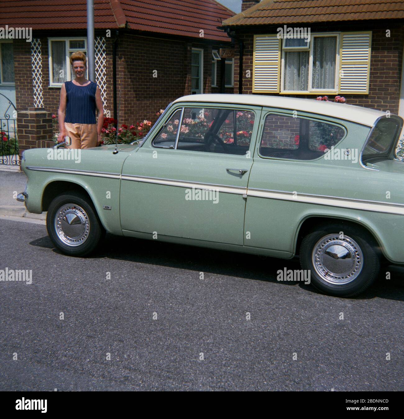 1970, historisch, außerhalb eines Bungalows in einer Vorstadtstraße, eine Besitzerin steht stolz neben ihrem zweitürigen Ford Anglia 105E-Wagen, einem beliebten Kleinfamilienauto, das zwischen 1959 und 1967 hergestellt wurde. Der 105E war das vierte Anglia-Modell und mit seinem neuen "amerikanisierten" Modell Unterschied sich von früheren Fahrzeugen mit abgerundeten Aufbauten und war ein großer Erfolg in Großbritannien mit Rekordverkäufen. Stockfoto