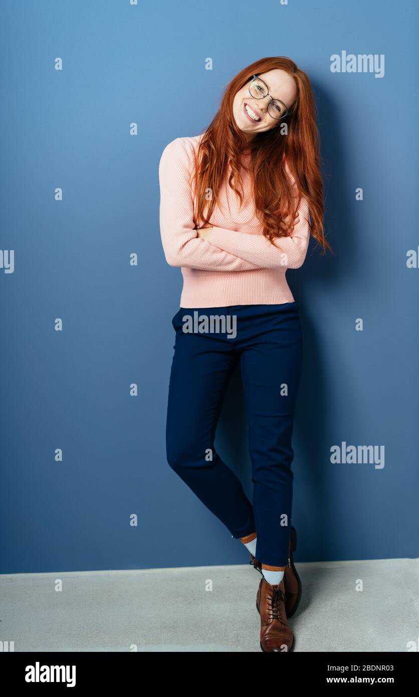 Entspannte trendige junge Rotschopf Frau in voller Länge posiert mit gefalteten Armen lächelnd an der Kamera gegen eine blaue Innenwand Stockfoto