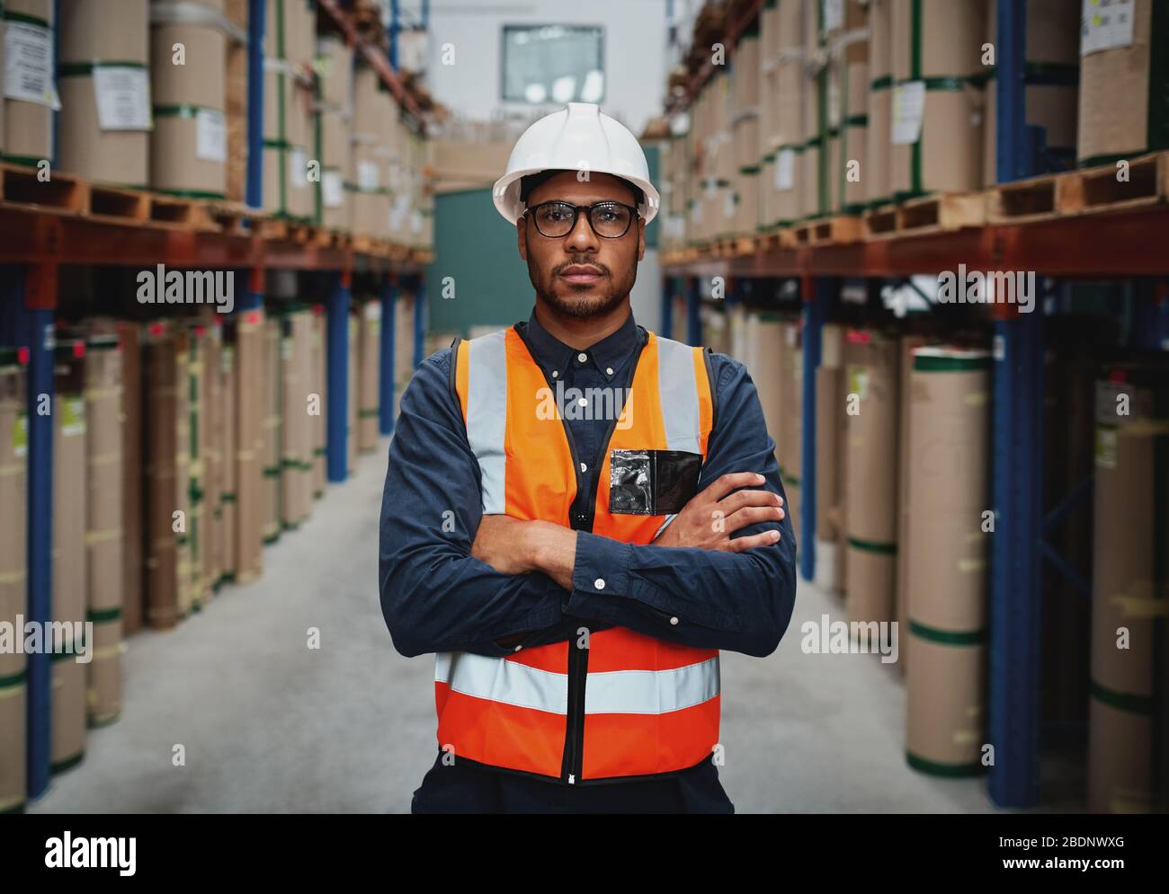 Selbstbewusster junger Manager mit gekreuzten Armen, die orangefarbene Weste tragen und weißer Hardhut in der Produktionseinheit, die auf die Kamera blickt Stockfoto