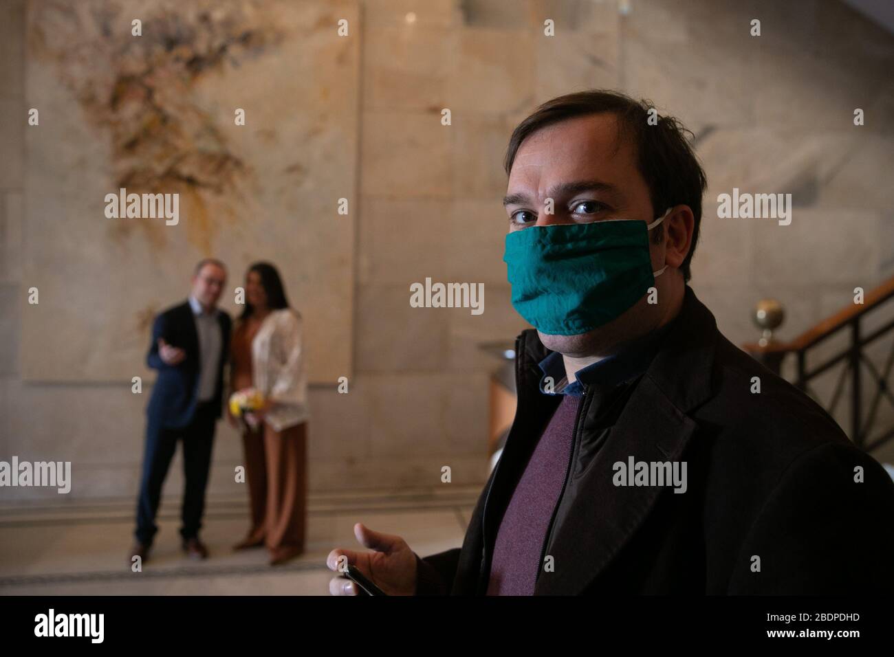 Athen, Griechenland. April 2020. Ein Hochzeitsgast, der eine chirurgische Maske trägt, blickt in die Kamera, da das bald darauf geborene Ehepaar am 8. April 2020 im Hintergrund in der Eingangslobby des Athener Rathauses in Athen, Griechenland, zu sehen ist. "Liebe herrscht" ist die Botschaft zweier entschiedener Paare, die vom Sperrverhängnis unbeirrt waren und am Mittwoch im Athener Rathaus geheiratet hatten. TO GO WITH'Feature: Liebe herrscht vor, wenn Paare in Griechenland den Knoten während der COVID-19-Sperrgutschrift binden: Lefteris Partsalis/Xinhua/Alamy Live News Stockfoto