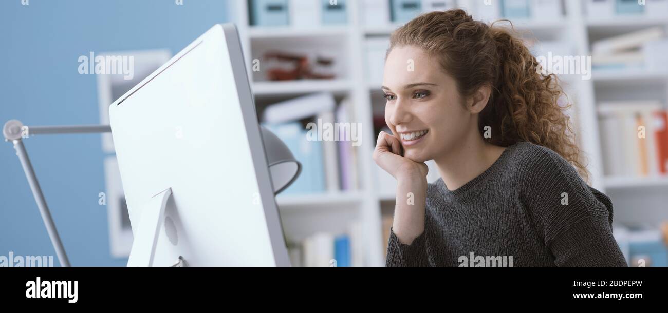 Junge lächelnde, schöne Frau, die einen Computer im Büro verwendet und online eine Verbindung herstellt, studiert und arbeitet Stockfoto