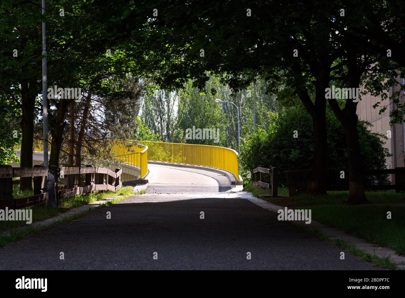 Gelbe Fußgängerbrücke über die Straße, die an sonnigen Sommertagen links vom Schatten der Laubbäume im Park abbiegt Stockfoto