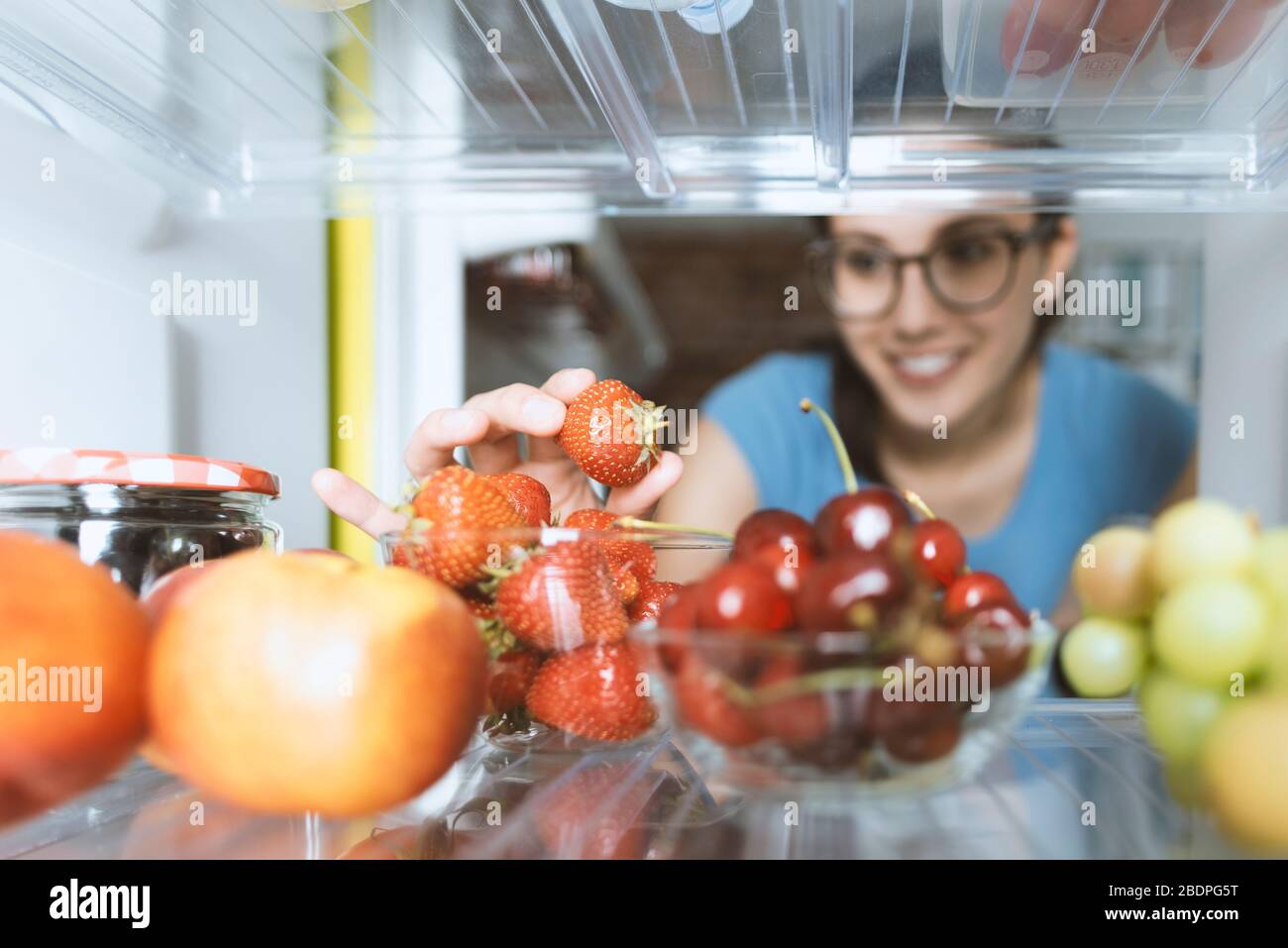 Junge lächelnde Frau, die frische Erdbeeren, den Kühlschrank und Lebensmittel in den Regalen isst Stockfoto