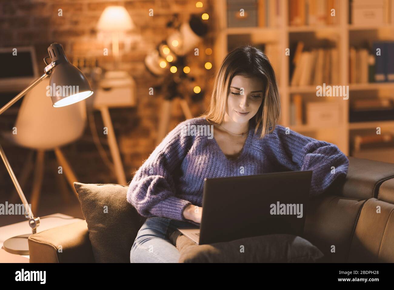 Junge attraktive Frau, die sich zu Hause auf dem Sofa entspannen und Filme online auf ihrem Laptop ansehen kann, Wohnzimmer im Hintergrund Stockfoto
