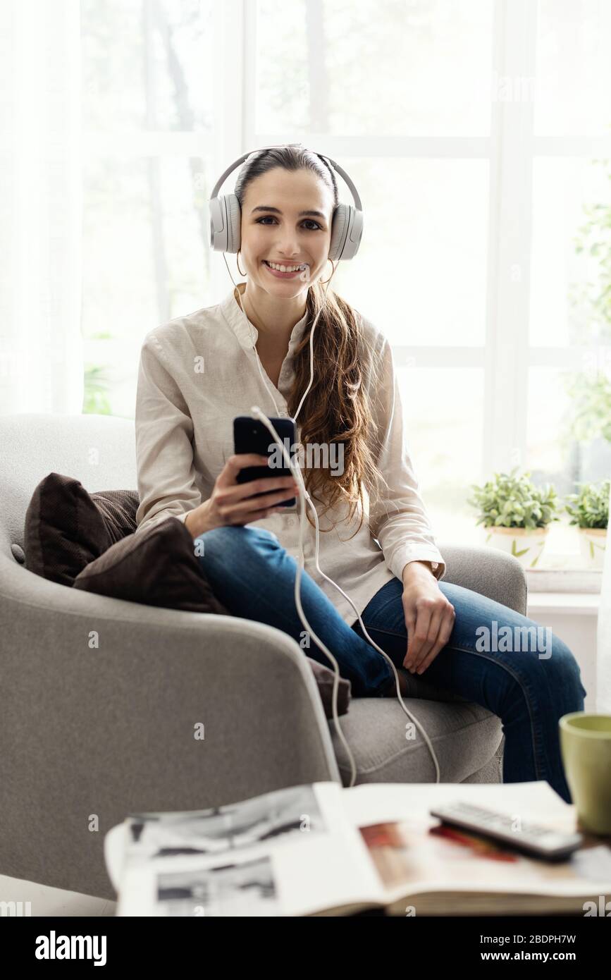 Junge lächelnde Frau, die zu Hause im Wohnzimmer auf dem Sessel sitzt und Musik über Kopfhörer und Smartphone hört Stockfoto