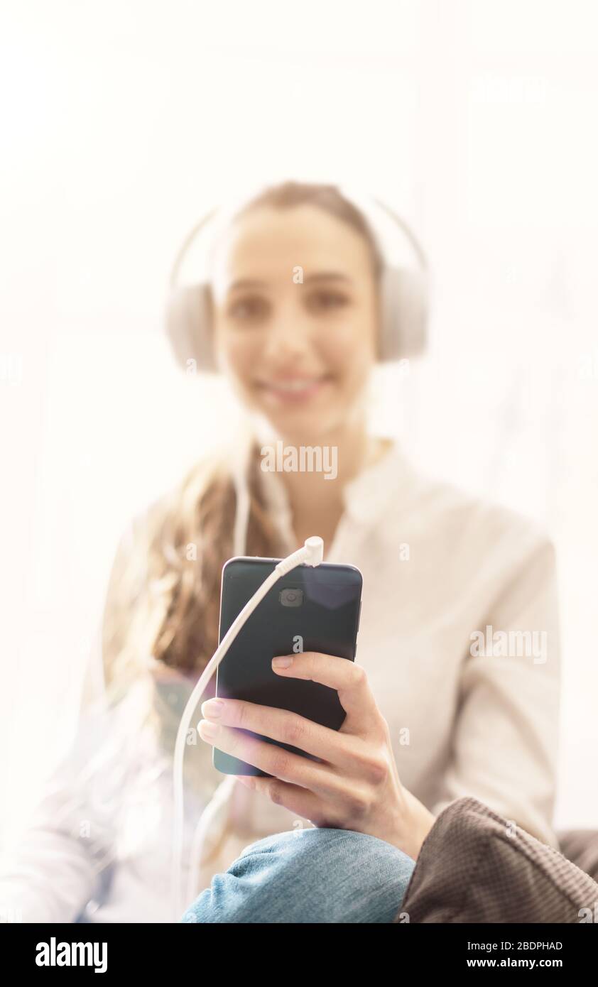 Junge lächelnde Frau, die zu Hause im Wohnzimmer auf dem Sessel sitzt und Musik über Kopfhörer und Smartphone hört Stockfoto