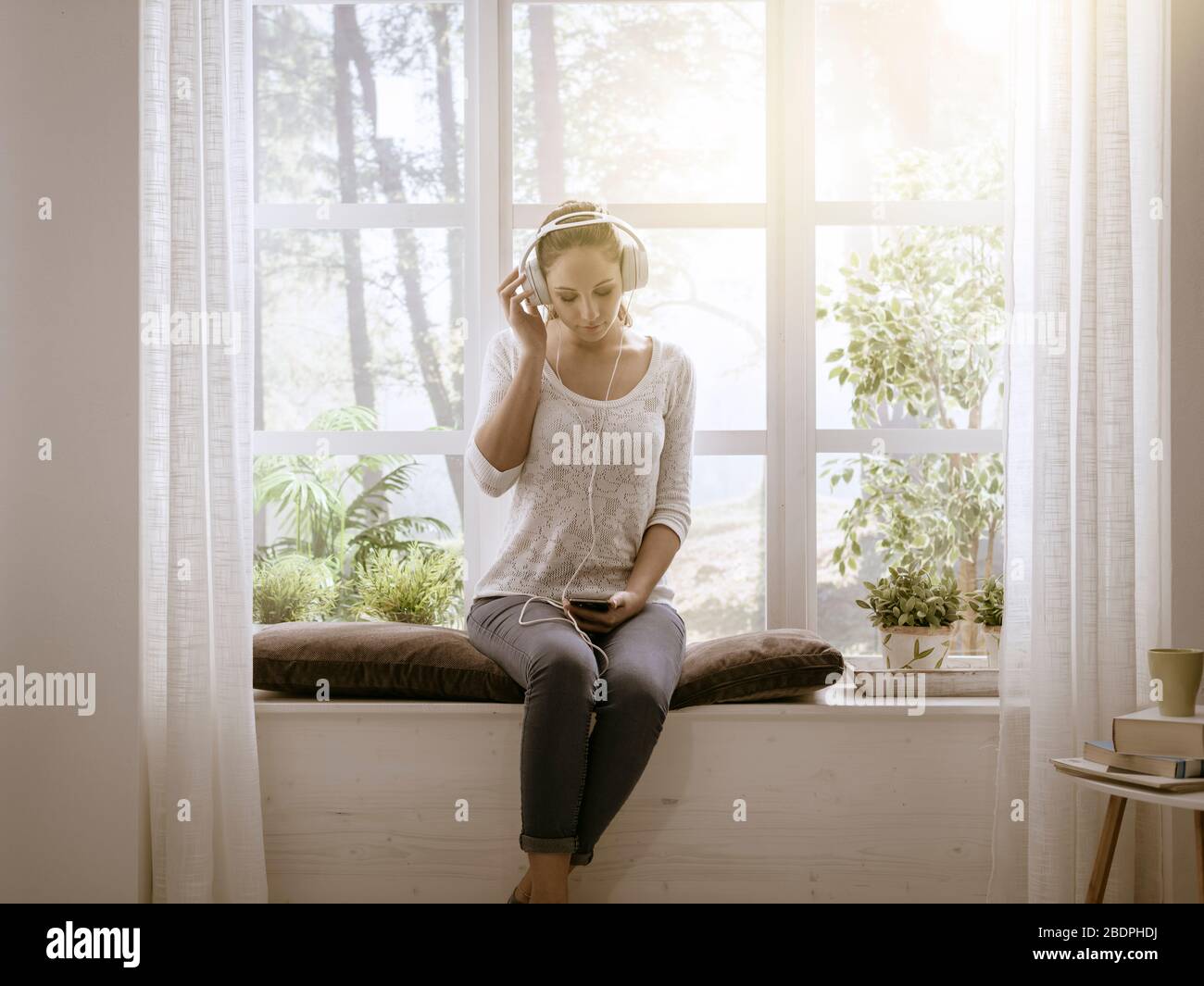 Entspannte glückliche Frau, die zu Hause neben einem Fenster sitzt und Musik über Kopfhörer hört Stockfoto