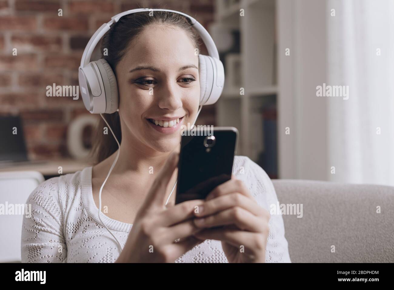 Fröhliche junge Frau, die zu Hause Musik hört, verbindet sie mit ihrem Smartphone und trägt Kopfhörer Stockfoto