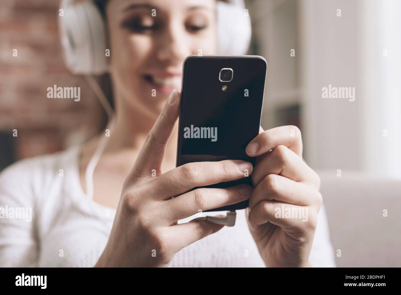Fröhliche junge Frau, die zu Hause Musik hört, verbindet sie mit ihrem Smartphone und trägt Kopfhörer Stockfoto