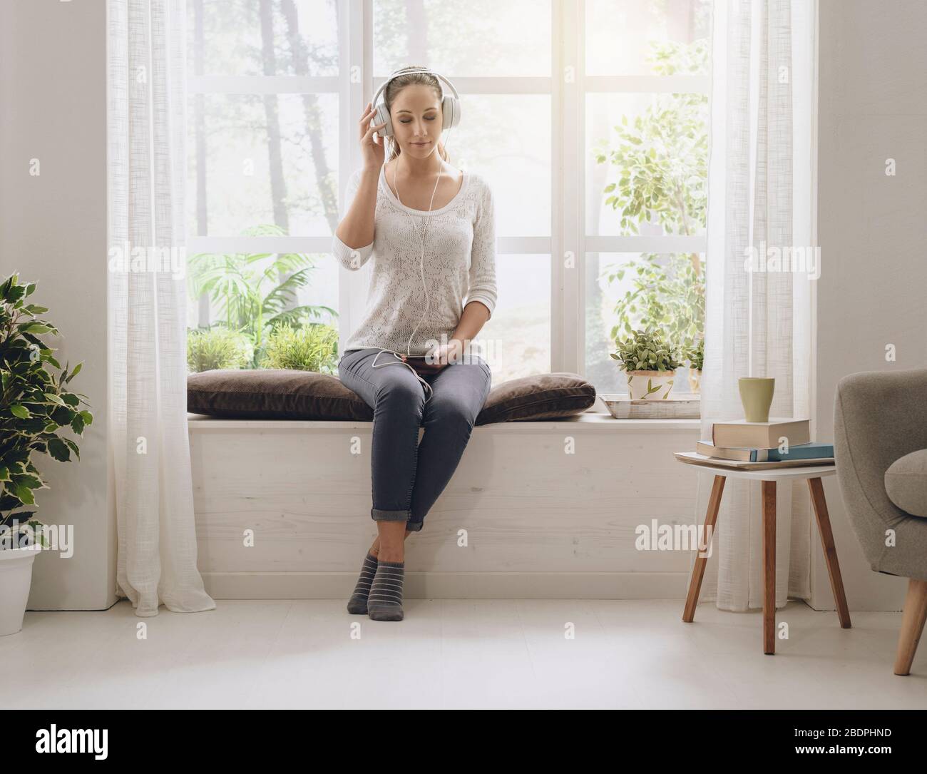 Entspannte glückliche Frau, die zu Hause neben einem Fenster sitzt und Musik über Kopfhörer hört Stockfoto