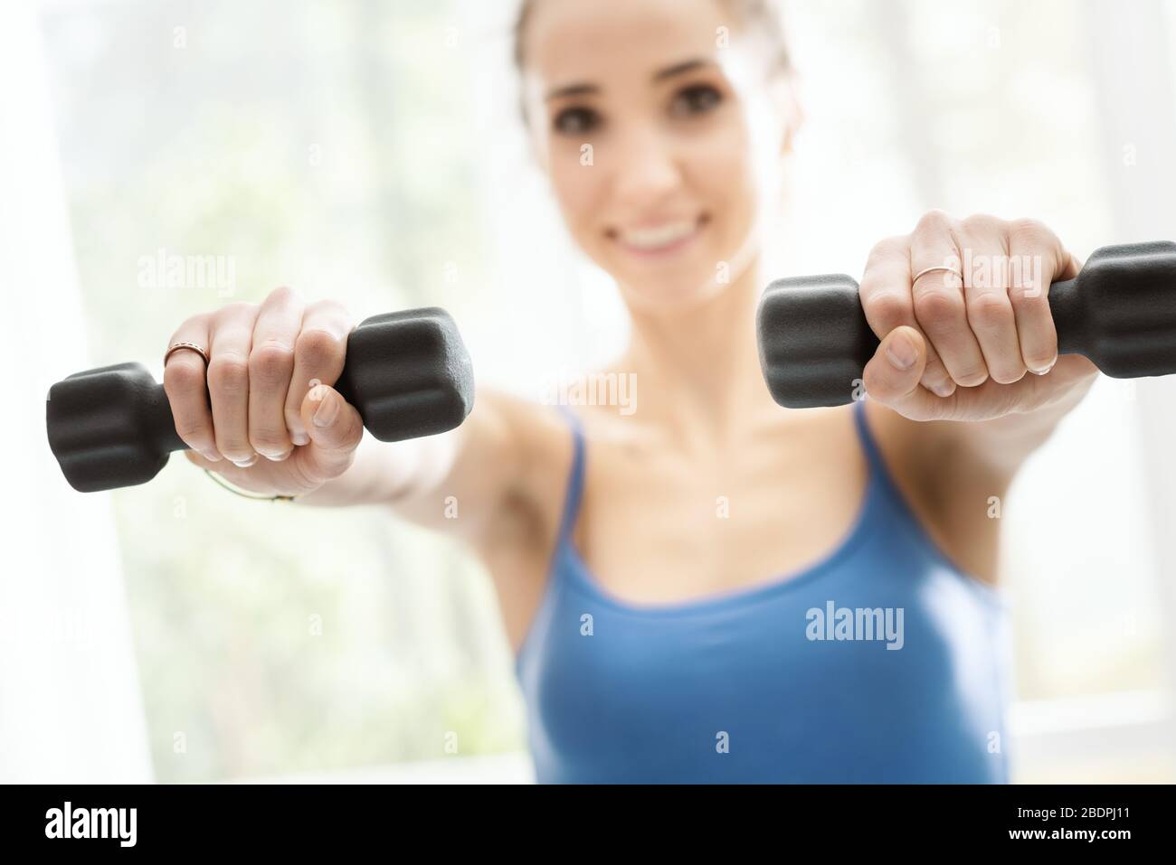 Junge lächelnde Frau, die zu Hause neben einem Fenster trainiert, hebt Kurzhanteln, Fitness- und Sportkonzept Stockfoto