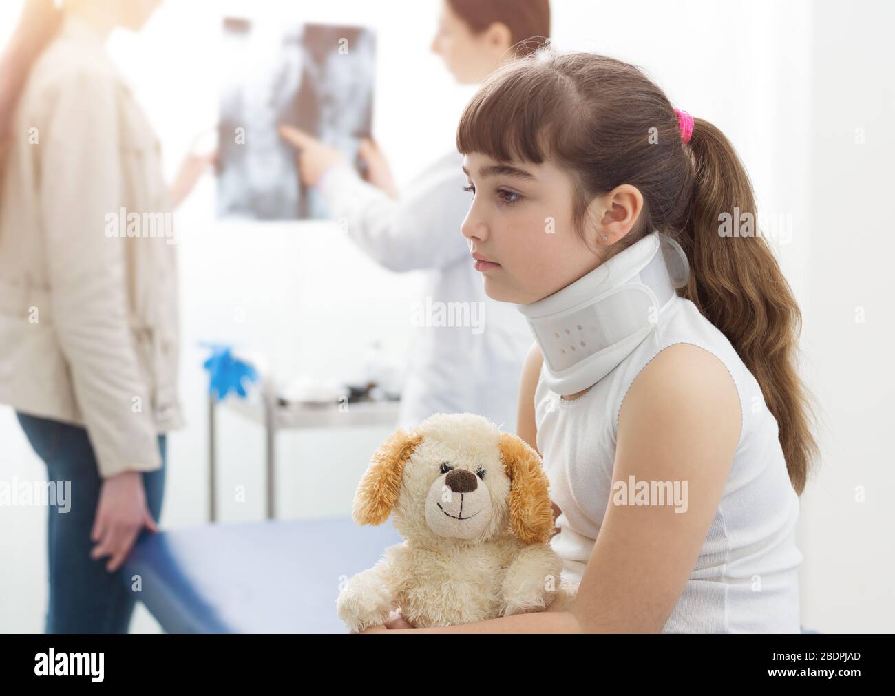 Mädchen mit Halsband in der Arztpraxis, sie hält ihren Teddybären, der Radiologe spricht im Hintergrund mit ihrer Mutter Stockfoto