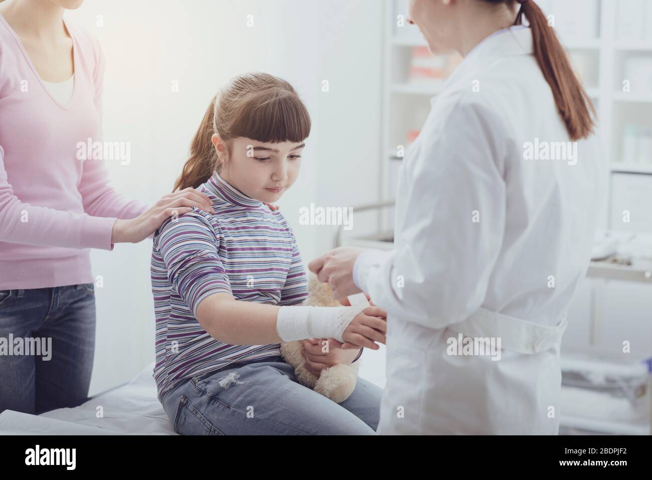 Doktor, der Gaze und Bandage auf das zerbrochene Handgelenk eines Mädchens wickelt, die Mutter steht neben der Tochter, erster Hilfe und Gesundheitskonzept Stockfoto