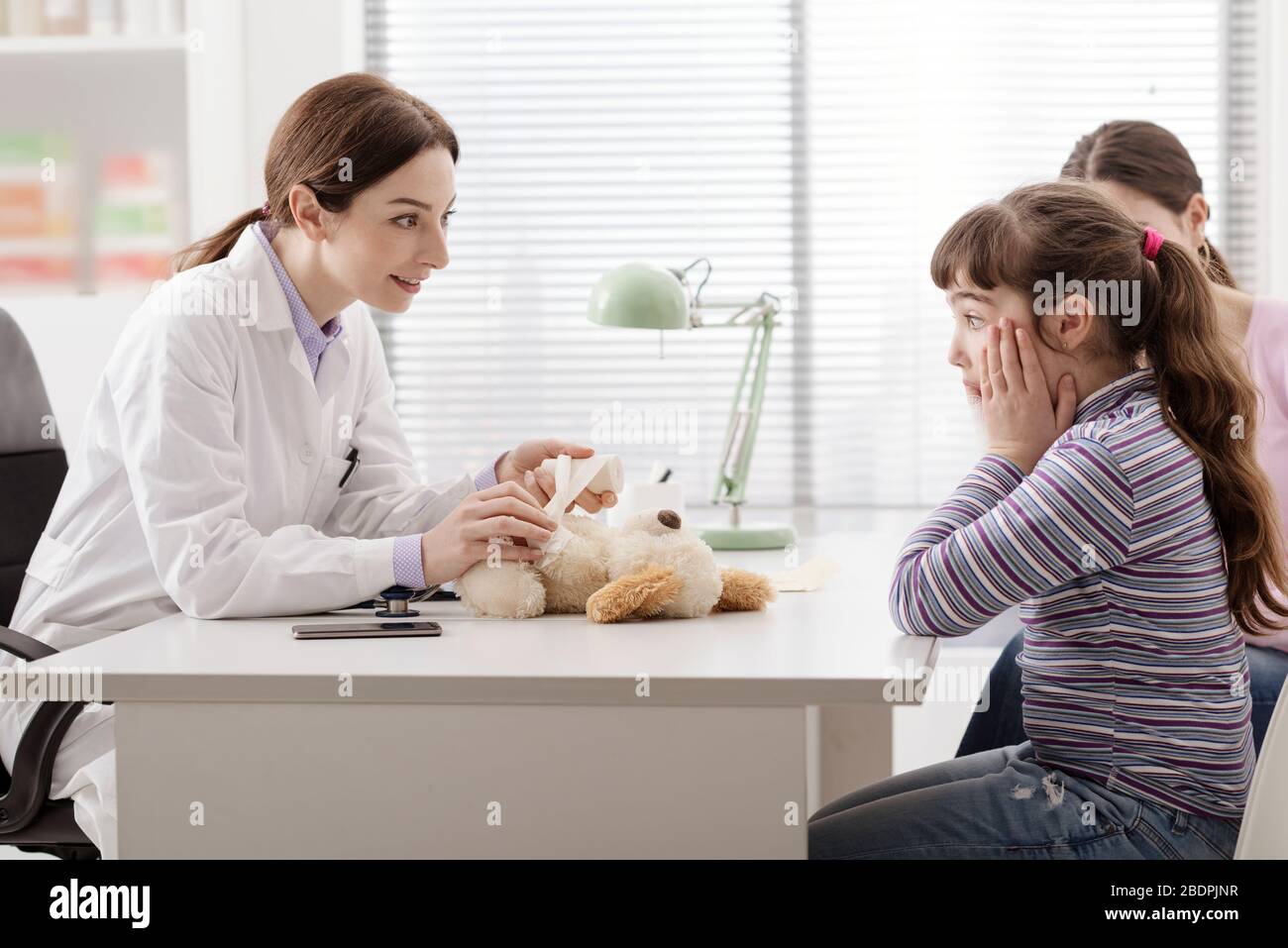 Verspielter Arzt, der den Teddybären eines Mädchens mit einem Verband umhüllt, ist das Mädchen schockiert, das Konzept von Gesundheit und Kindern Stockfoto