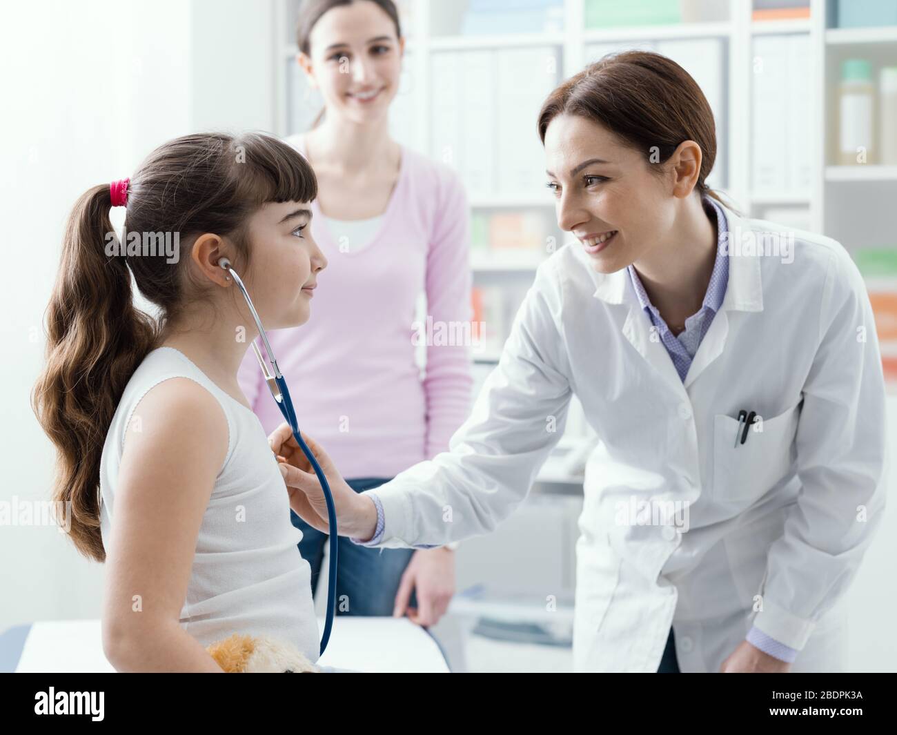 Weibliche Ärztin untersucht ein süßes lächelndes Mädchen mit einem Stethoskop, die Mutter ist neben ihr, Kinder- und Gesundheitskonzept Stockfoto