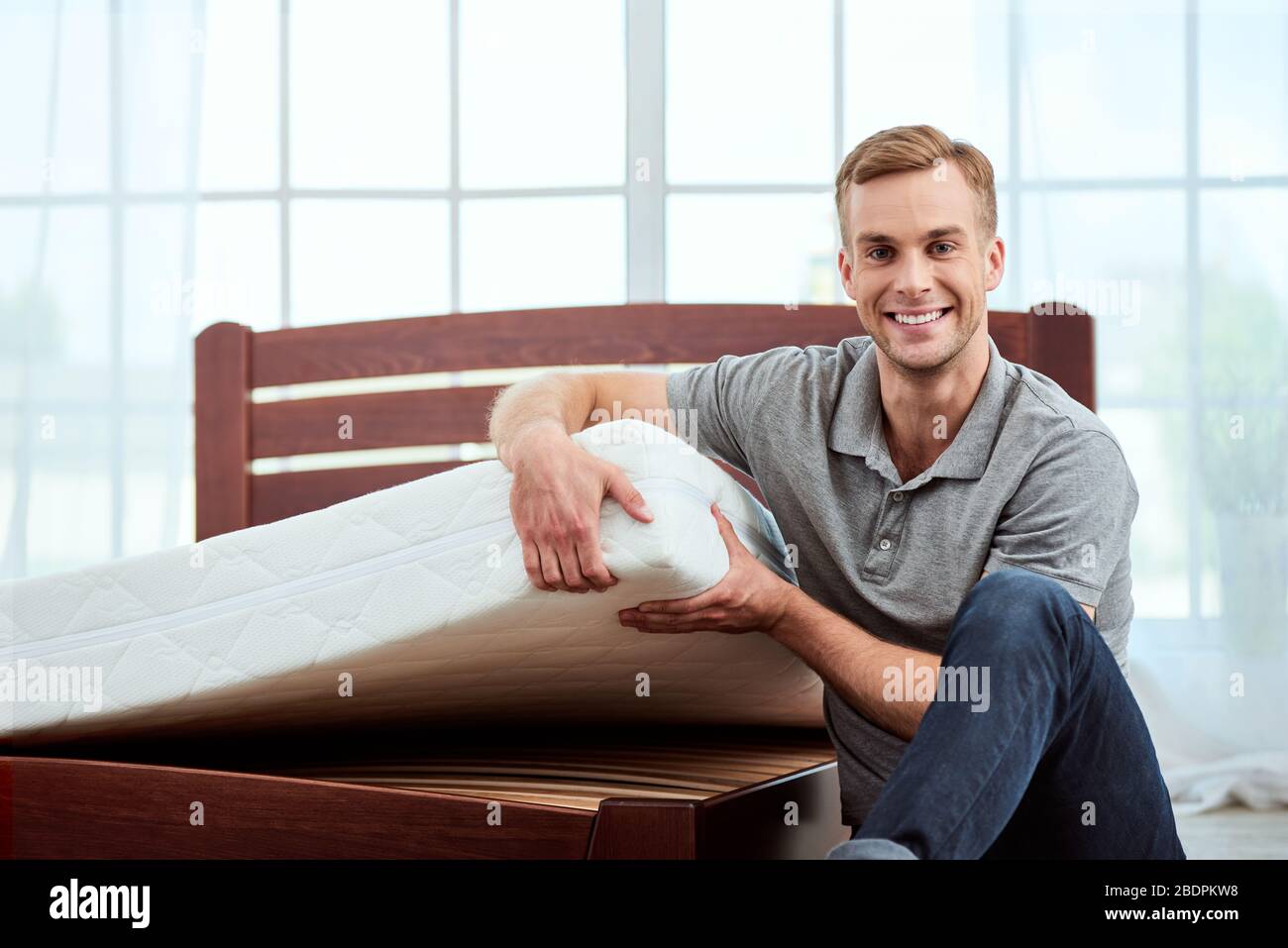 Junge und glückliche gutaussehende Mann zeigt große und komfortable orthopädische Matratze vor der Kamera und lächelnd. Schlafzimmer. Startseite. Gesundheitswesen Stockfoto