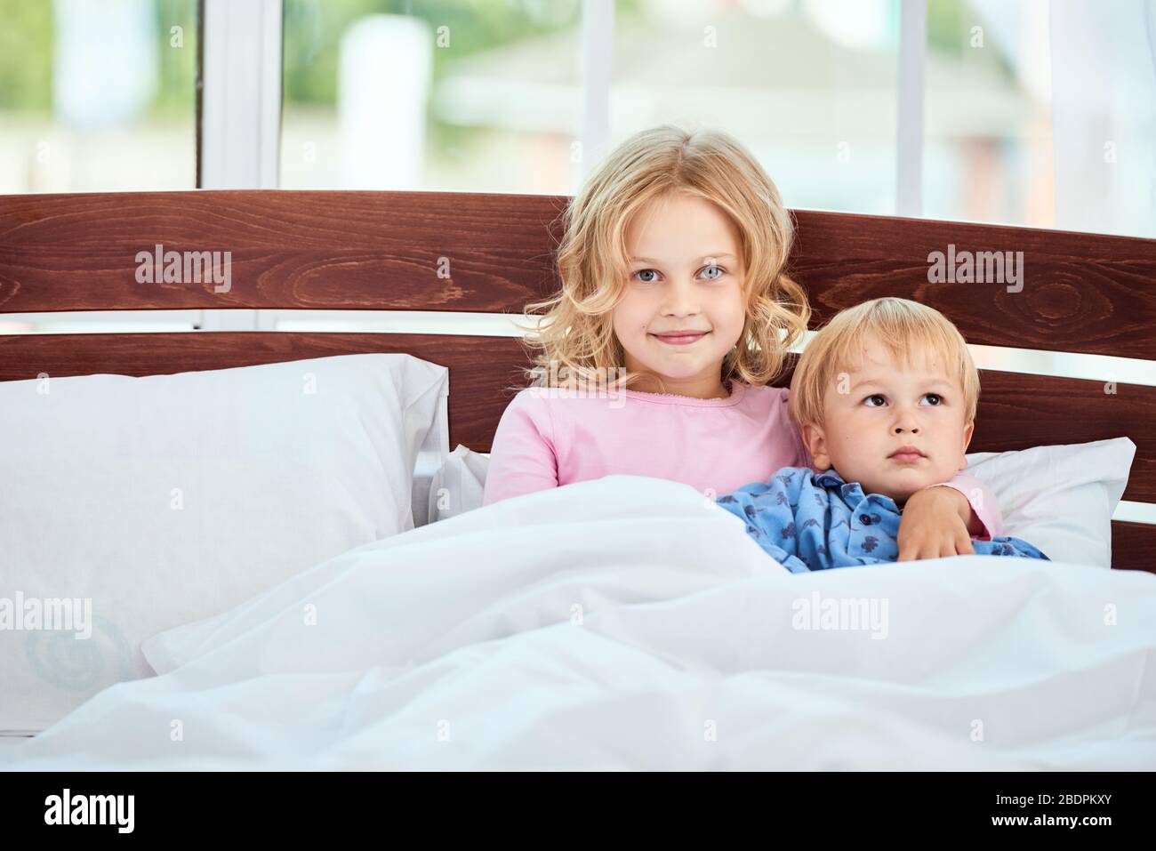 Portrait der Heilung kleine Schwester und Bruder im Pyjama Blick auf Kamera und lächelnd, während zusammen in einem großen Bett zu Hause liegen. Quarantäne. Zeit zusammen verbringen. Familie Stockfoto