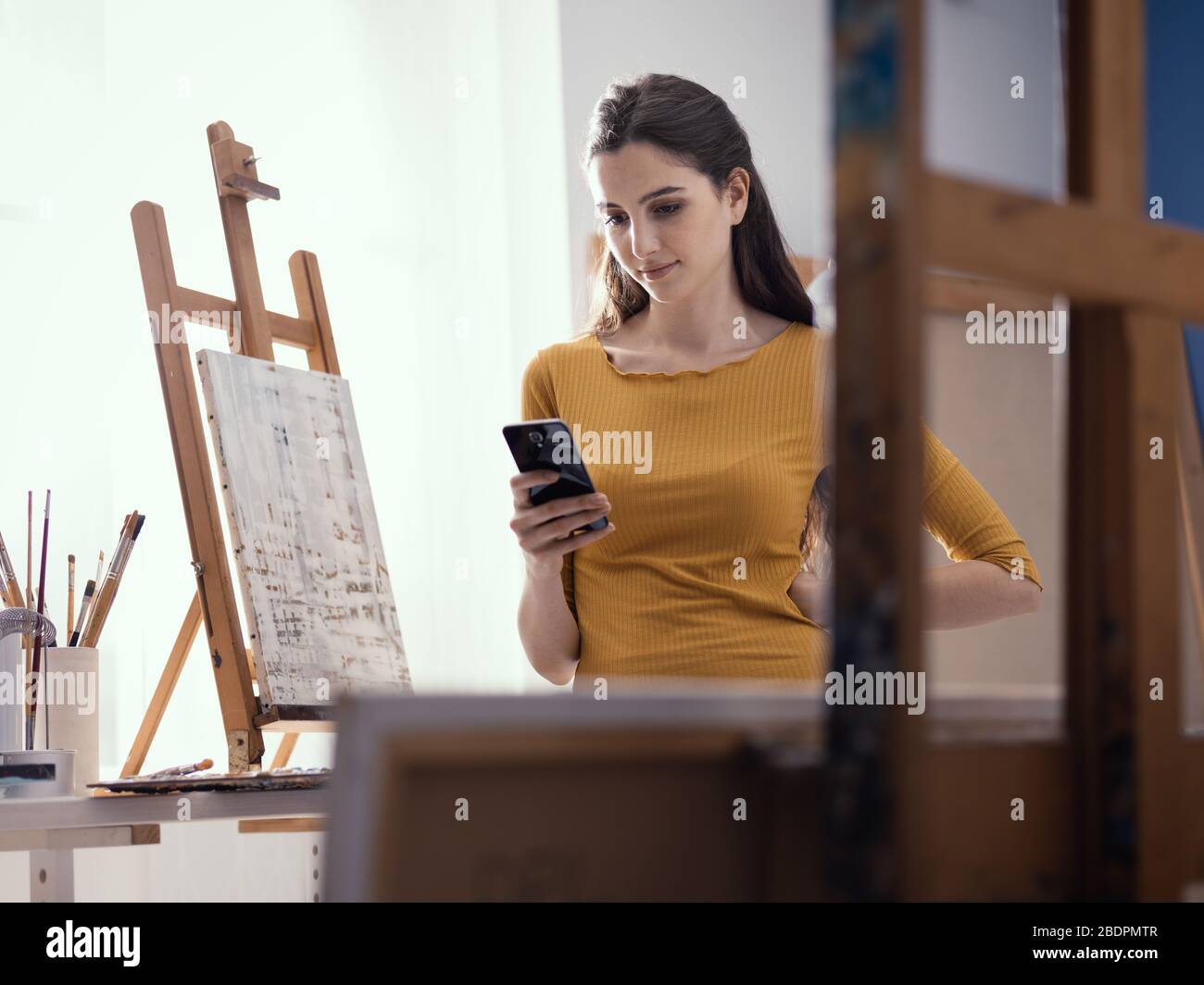 Fröhliche junge Künstlerin in ihrem Kunststudio steht sie neben ihrer Malerei und plaudert mit ihrem Smartphone Stockfoto