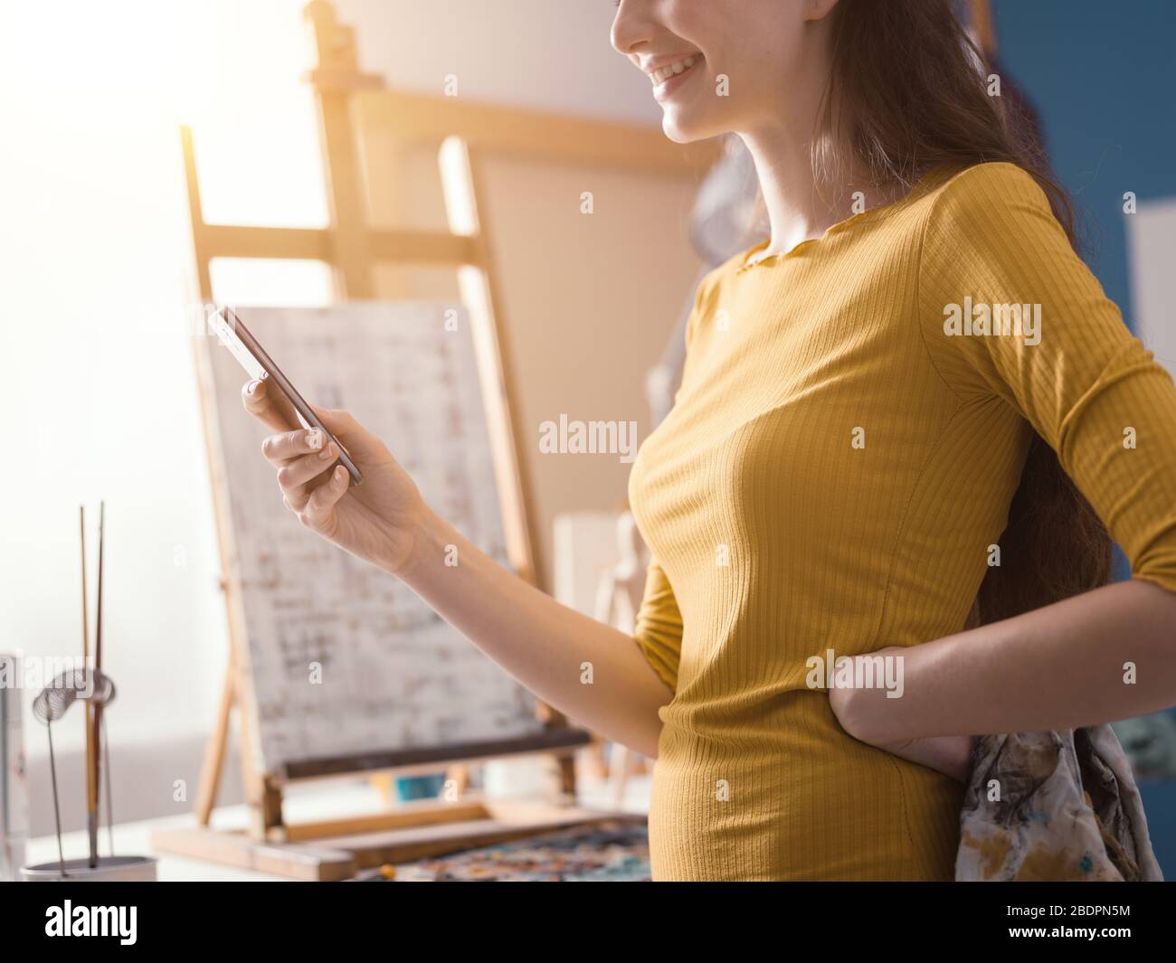 Fröhliche junge Künstlerin in ihrem Kunststudio steht sie neben ihrer Malerei und plaudert mit ihrem Smartphone Stockfoto