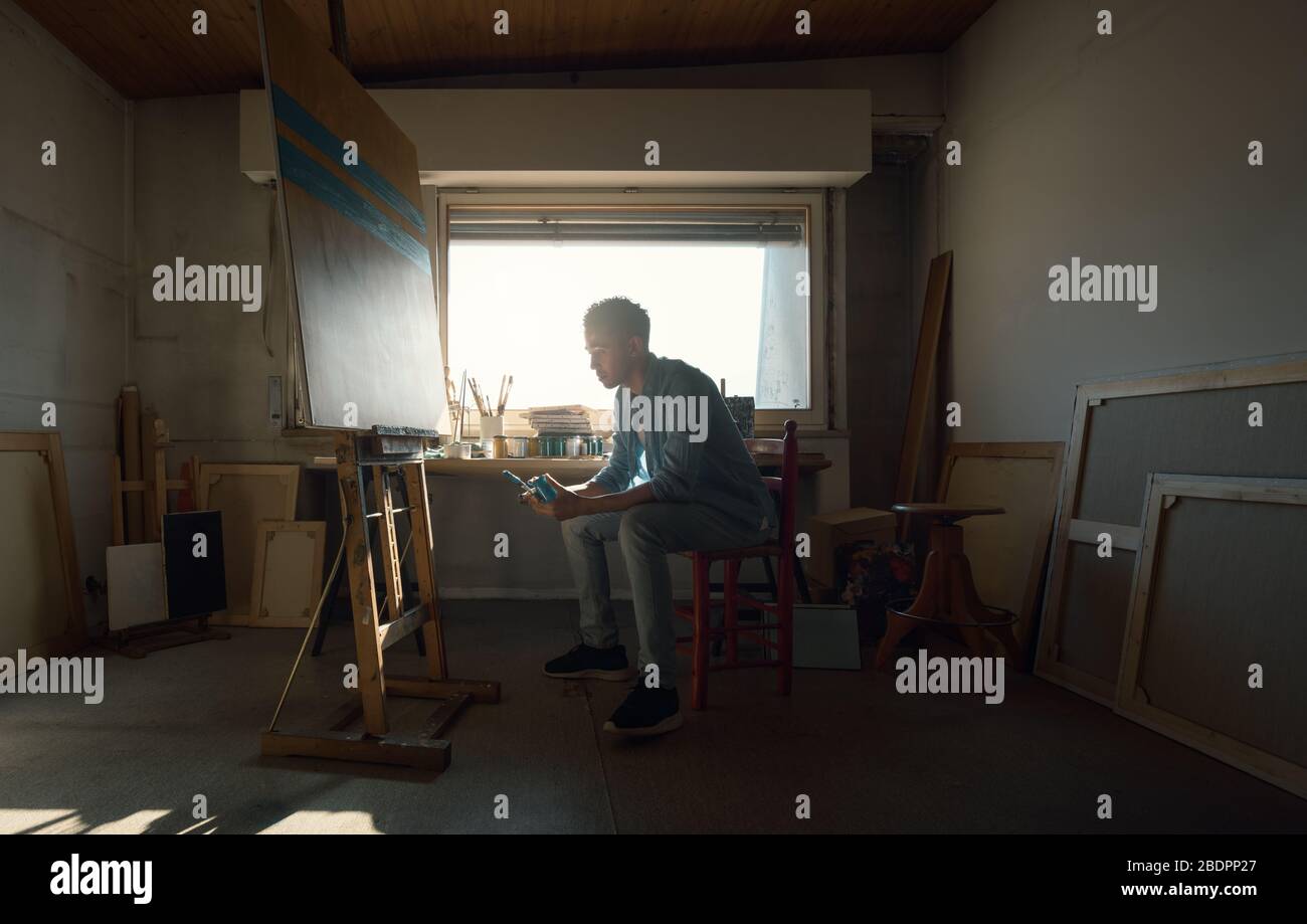 Kreativer junger Künstler, der in seinem Atelier sitzt und an seinem Gemälde arbeitet: Er sitzt, hält einen Pinsel und einen Krug mit Farbe Stockfoto