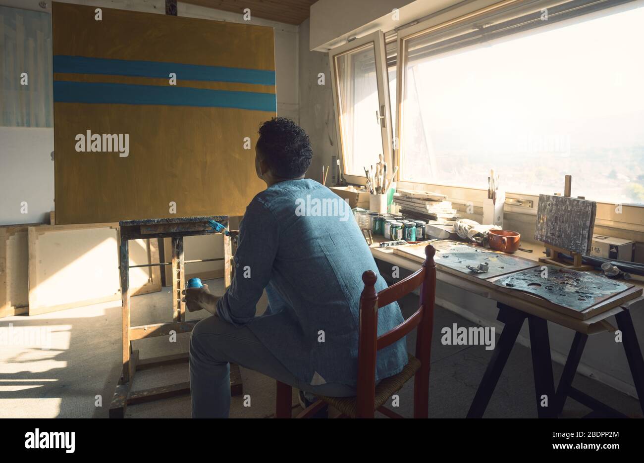 Kreativer junger Künstler, der in seinem Atelier sitzt und an seinem Gemälde arbeitet: Er sitzt, hält einen Pinsel und einen Krug mit Farbe Stockfoto