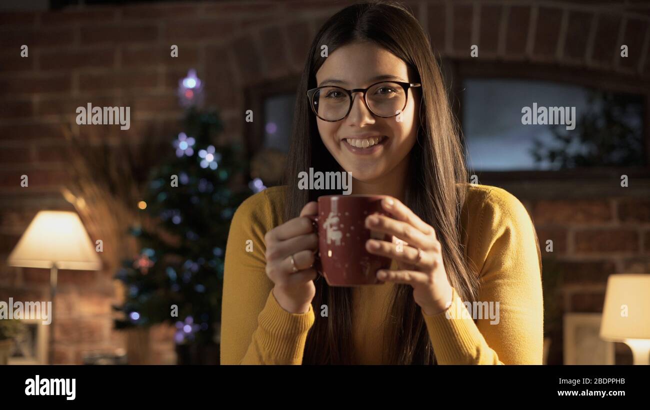 Fröhliche junge Frau, die zu Hause eine Tasse Tee hat, Weihnachtsbaum im Hintergrund, Weihnachtskonzept Stockfoto